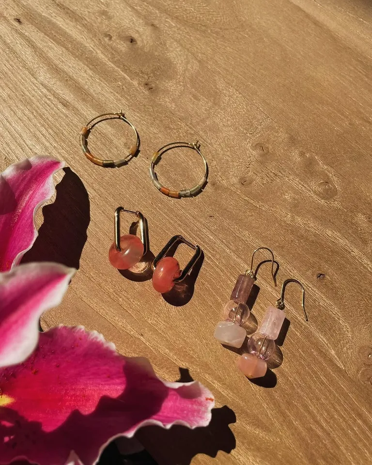 Three pairs of earrings with coloured stones on a wooden table, beside a pink lily